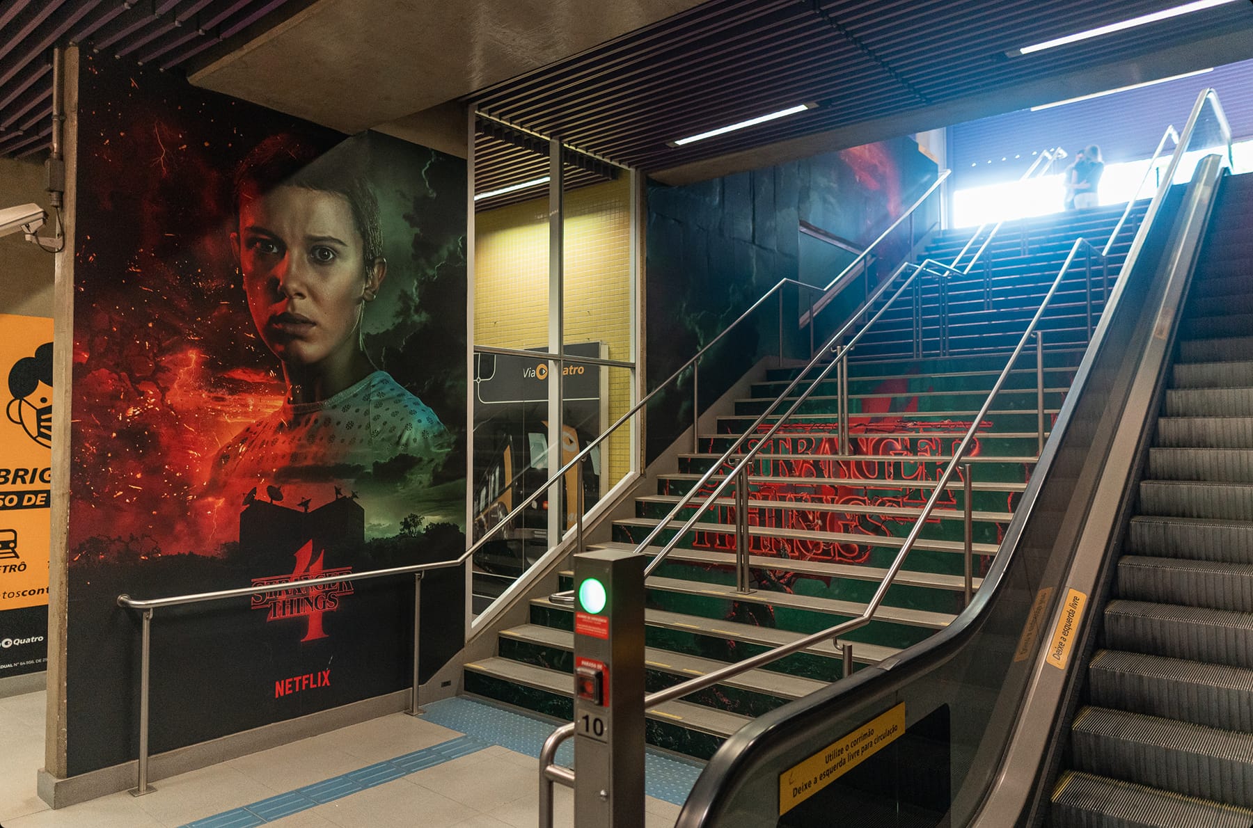 Estação de metrô com um mural de Stranger Things: o rosto da personagem Onze entre chamas vermelhas e mata escura, e o letreiro da série impresso nos degraus da escada ao lado da escada rolante