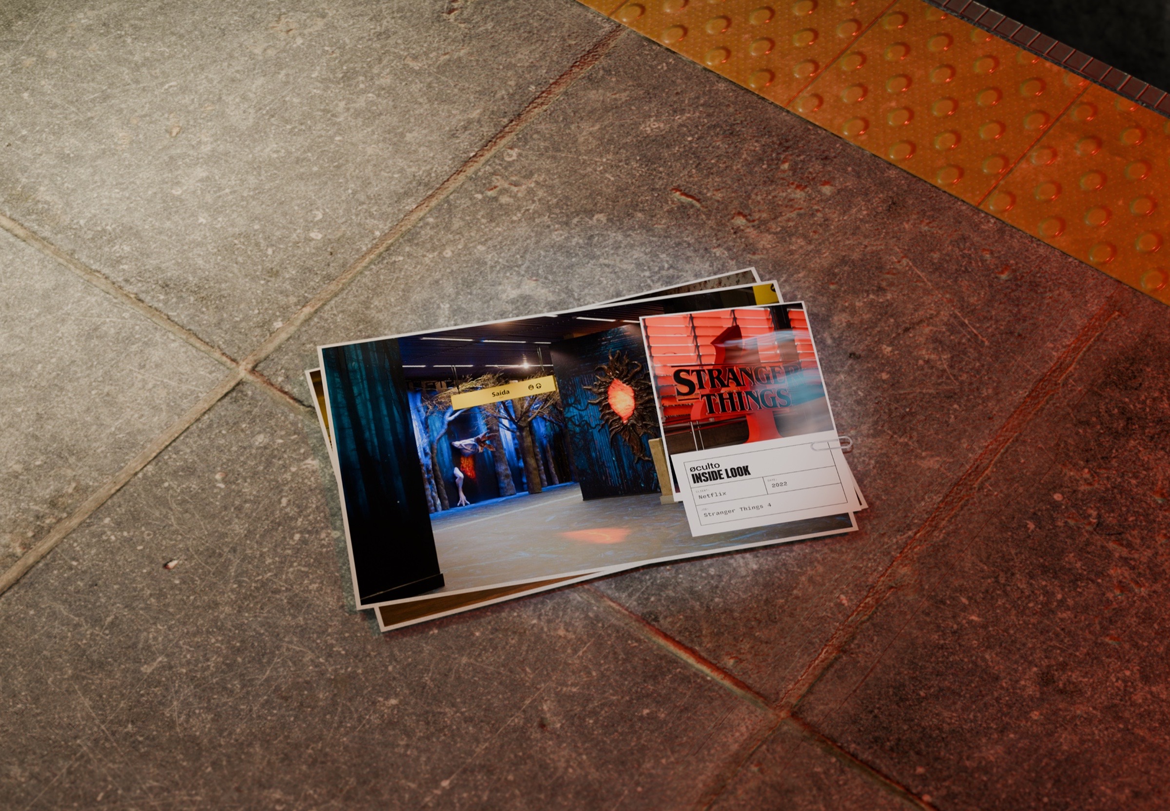 Printed photographs of the project stacked on the station's concrete floor beside the orange tactile strip, with an øculto Inside Look card identifying Netflix, Stranger Things 4, 2022