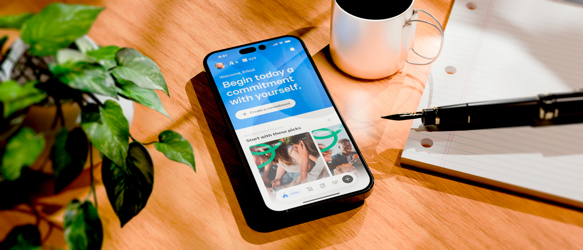 Phone on a wooden desk beside a coffee cup, a plant and a notebook with a fountain pen, showing the Alltagshelden home screen: Begin today a commitment with yourself, the create-a-commitment button and a first row of suggestions