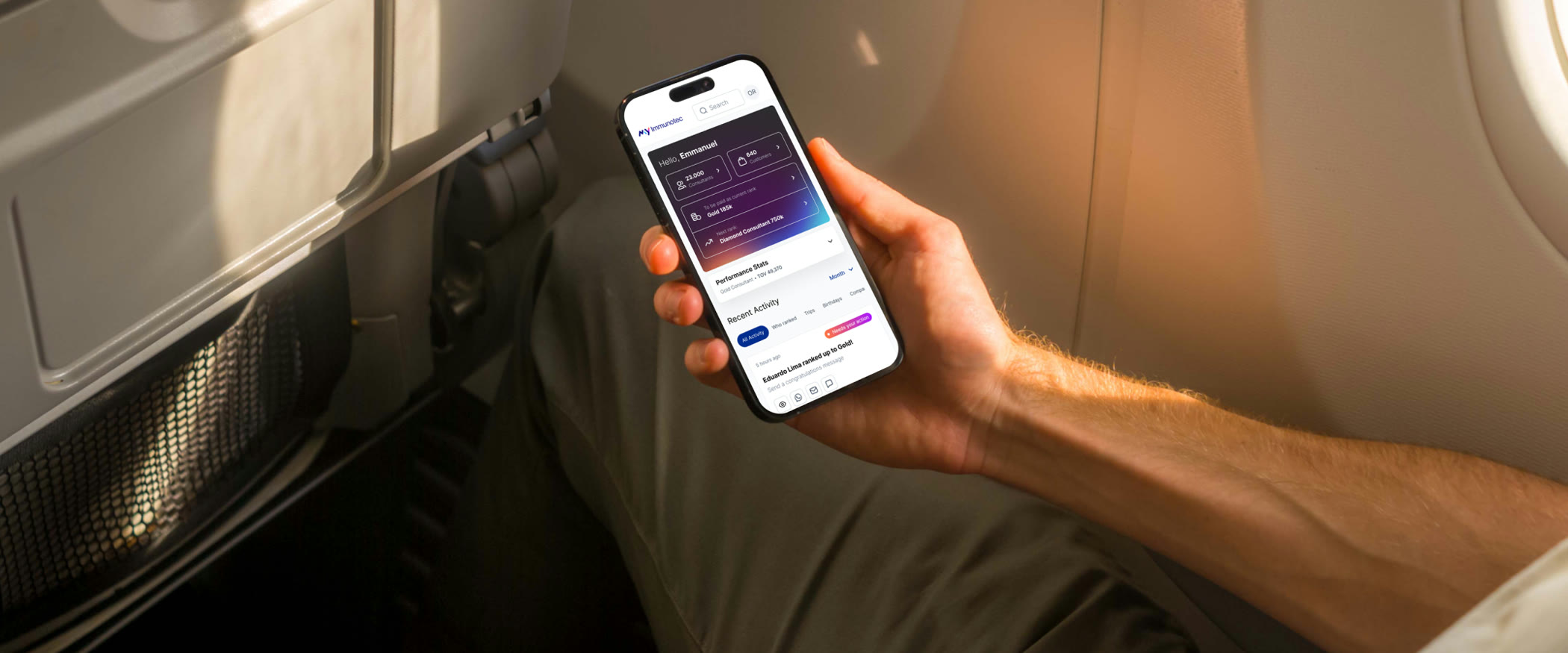 A man seated on a plane by the window, holding his phone with the MyImmunotec dashboard open: the greeting, the consultant and customer counts, his current rank and the network's recent activity