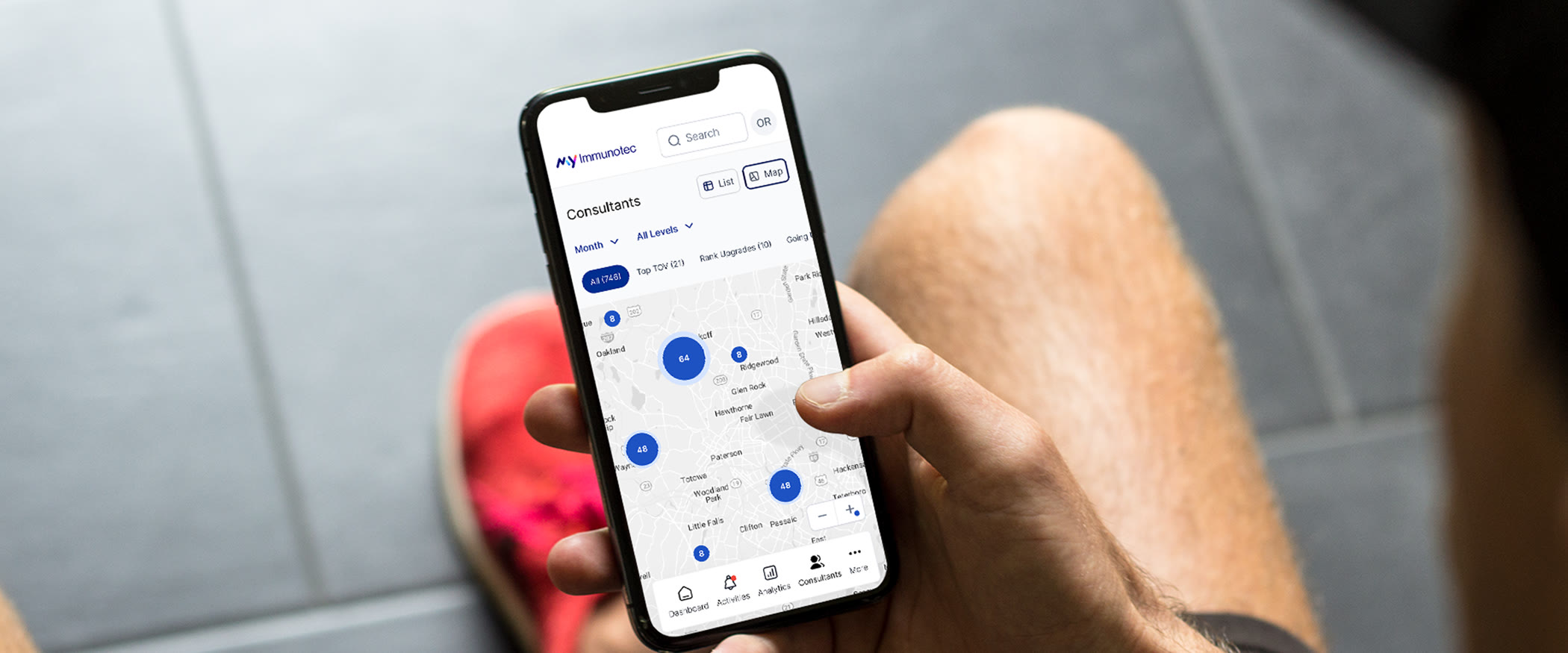 A hand holding a phone over a concrete floor, the MyImmunotec consultants screen open on the map view with the network's numbered clusters spread across the region