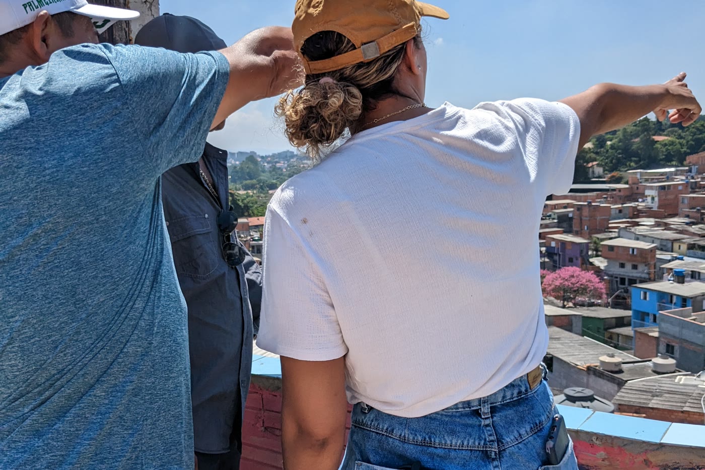 Two people seen from behind at a high point in a peripheral community, one of them pointing out across the valley of houses and rooftops opening up ahead, on a clear day