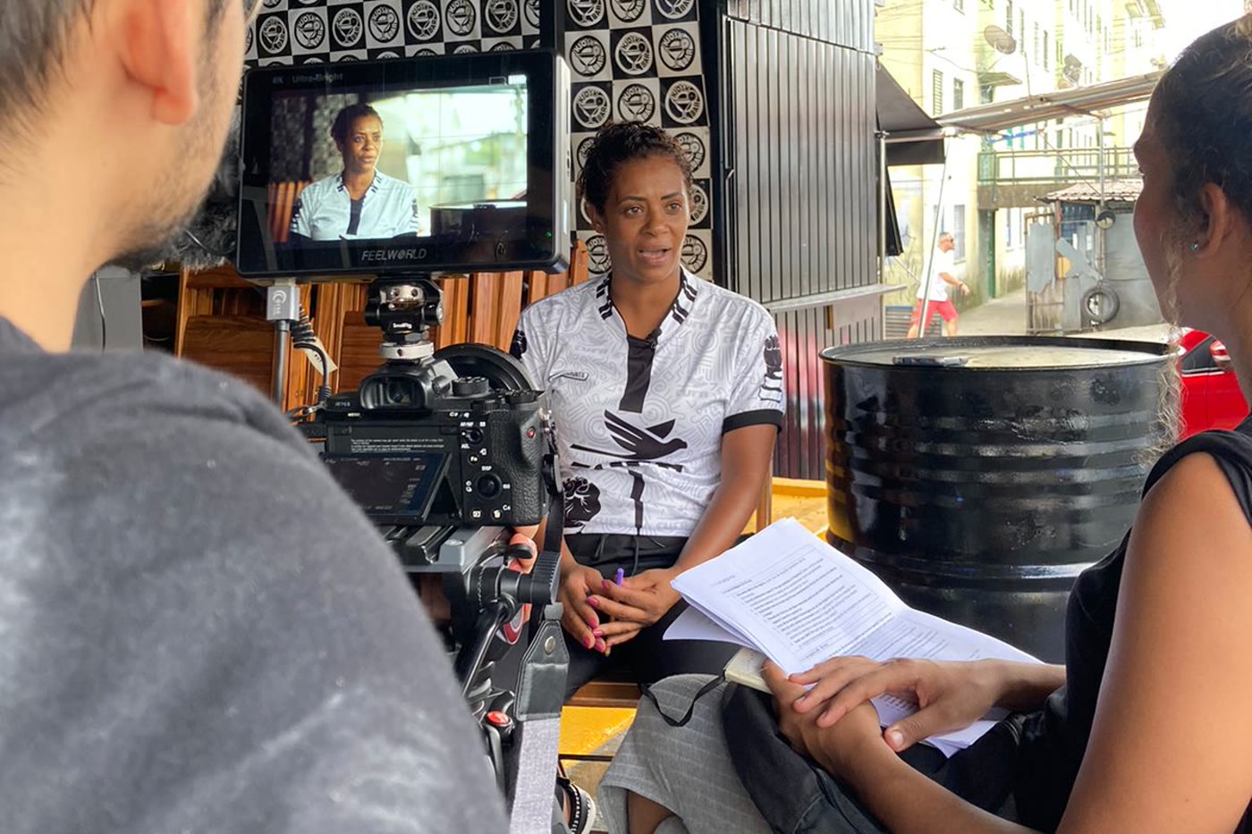 An interview being recorded on the street: the camera operator in the foreground, the business owner seated and answering, and the researcher holding the script, with the monitor showing the framing