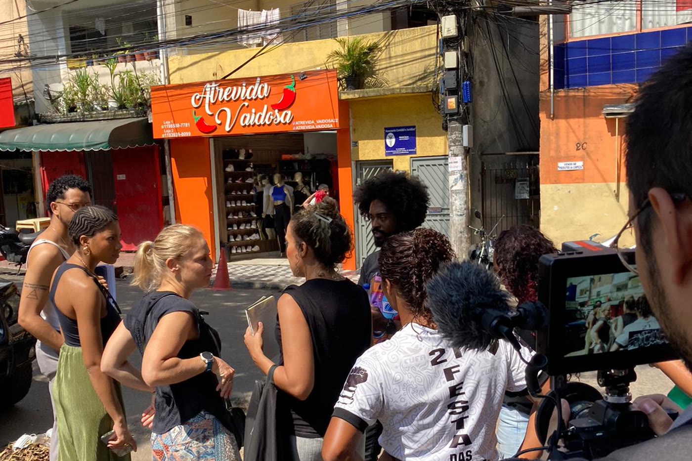 A group gathered on the pavement in front of a clothing shop, talking while the team records, with the street's shopfronts and power lines behind them
