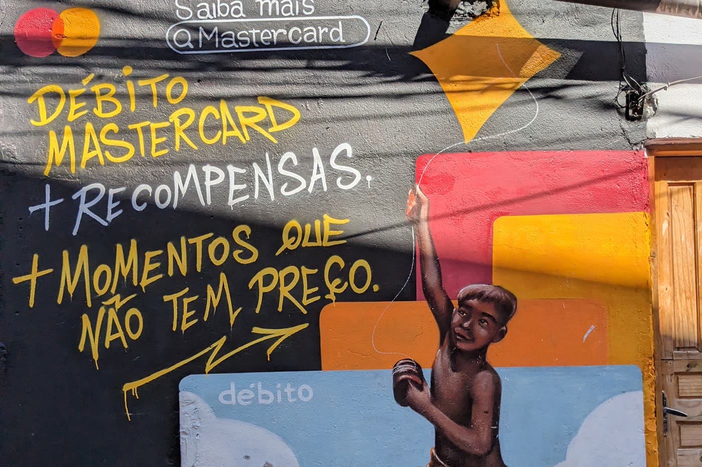 A mural painted on a wall in a peripheral community carrying the Mastercard brand, the lines Débito Mastercard plus rewards and moments that are priceless, and the figure of a child flying a kite