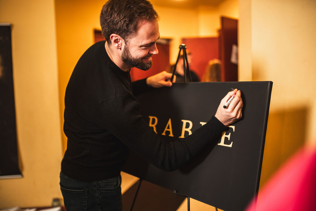 A man leaning over a black board carrying the Parable Studios logotype in gold, his hand on the letters, in a warm-lit room before the debut