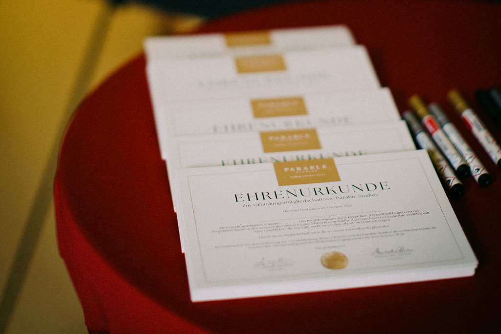 A stack of Parable Studios founding-membership certificates on a red table, each carrying the mark's gold seal, beside pens for the signatures