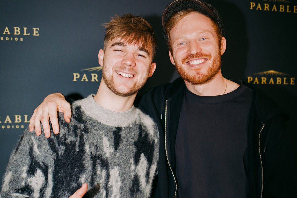 Two guests with arms around each other, smiling in front of the launch backdrop, the Parable Studios mark repeated in gold across the dark ground