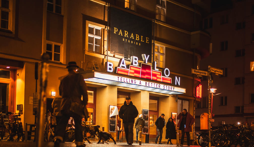 The Kino Babylon facade at night, the Parable Studios sign above the lit cinema marquee and the line Telling a Better Story, with people crossing the pavement